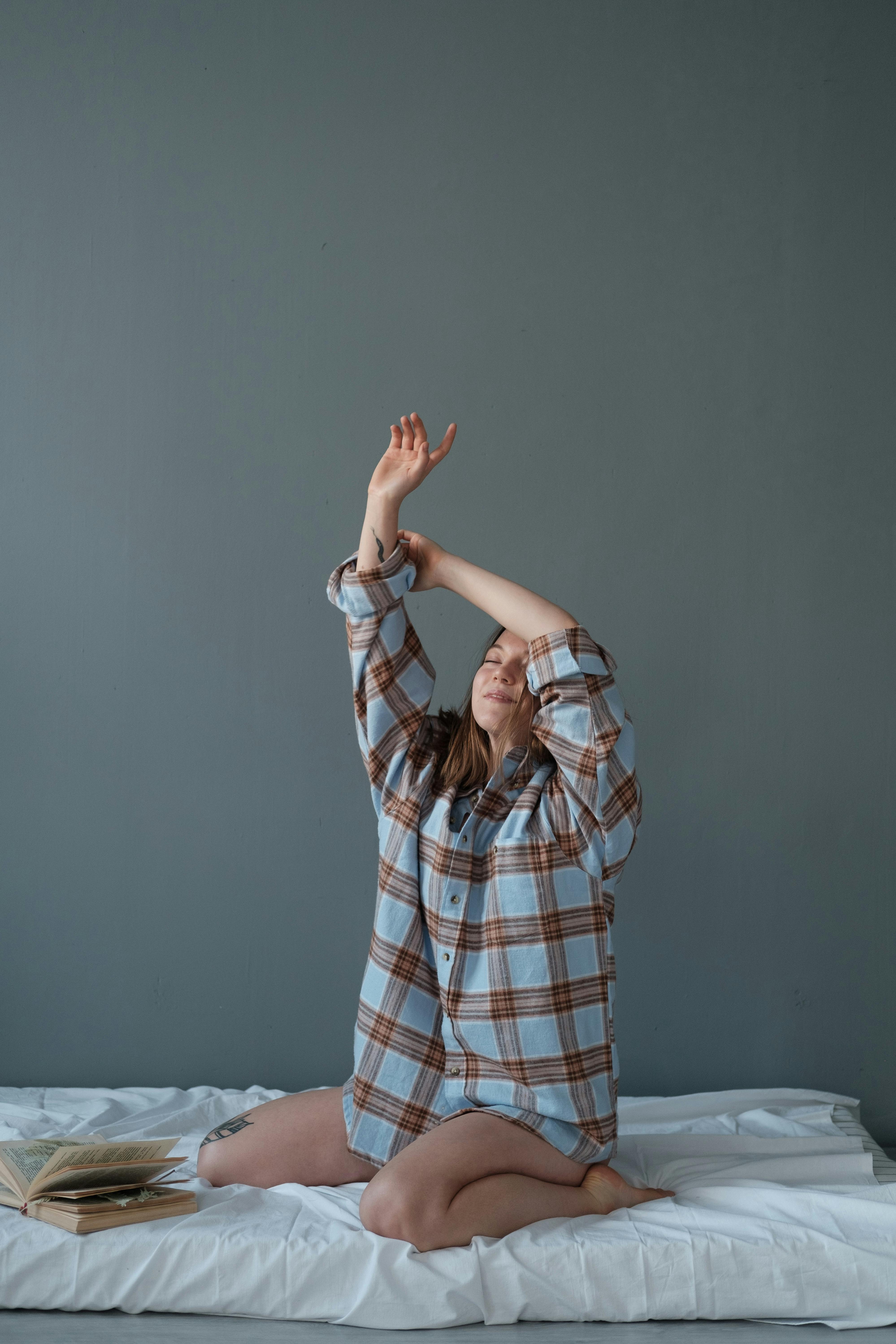 Woman in a plaid shirt kneeling and stretching on a bed indoors.