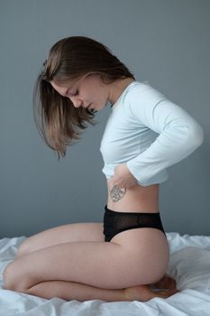 Side view of young female with tattoo sitting on bedsheet and looking at bare tummy