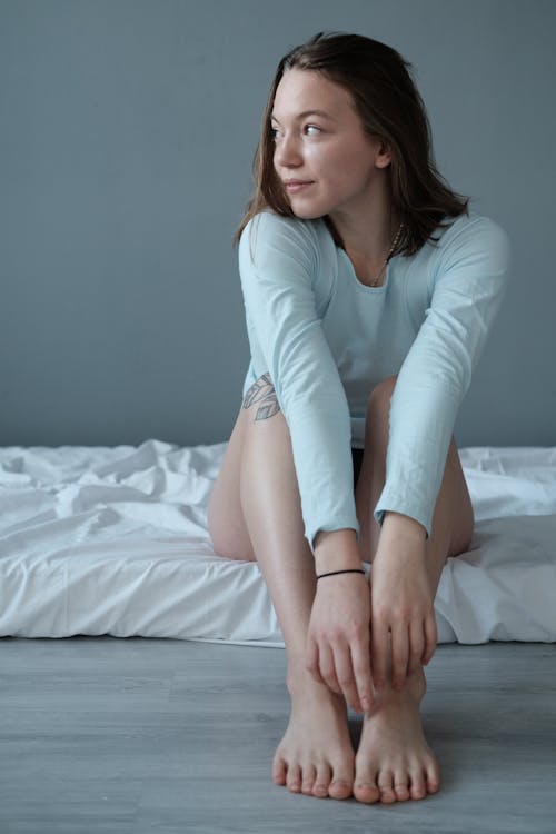 Full body of barefoot dreamy female in sleepwear sitting on bedsheet and looking away with gentle smile