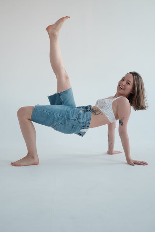 Full body side view of barefoot glad female wearing denim shorts and brasserie doing Reverse Table Top posture with hands raised and laughing joyfully