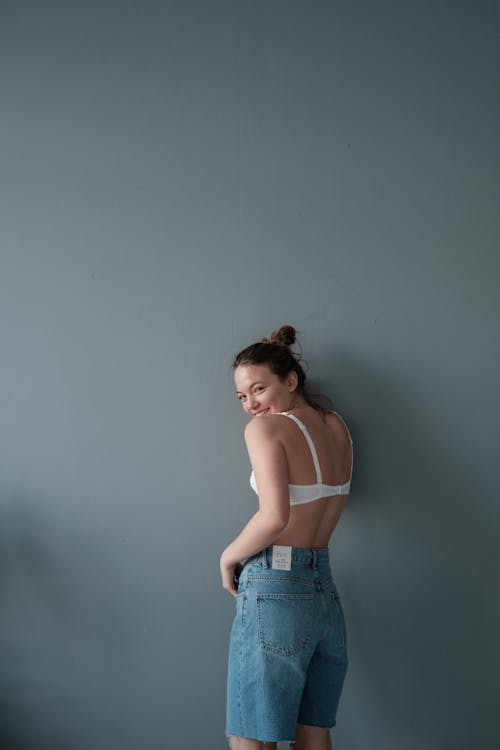 Side view of positive young female wearing denim shorts and white bra looking over shoulder at camera while standing near gray background