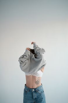 Anonymous female with tattoo in white bra putting on trendy gray hoodie while standing on white background in light studio