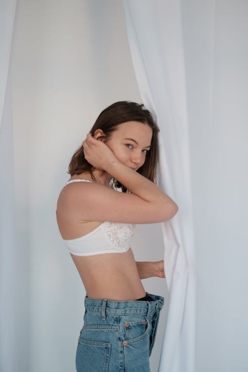 Side view of content young female wearing denim jeans and underwear tucking hair behind ear and looking at camera while standing near white curtains