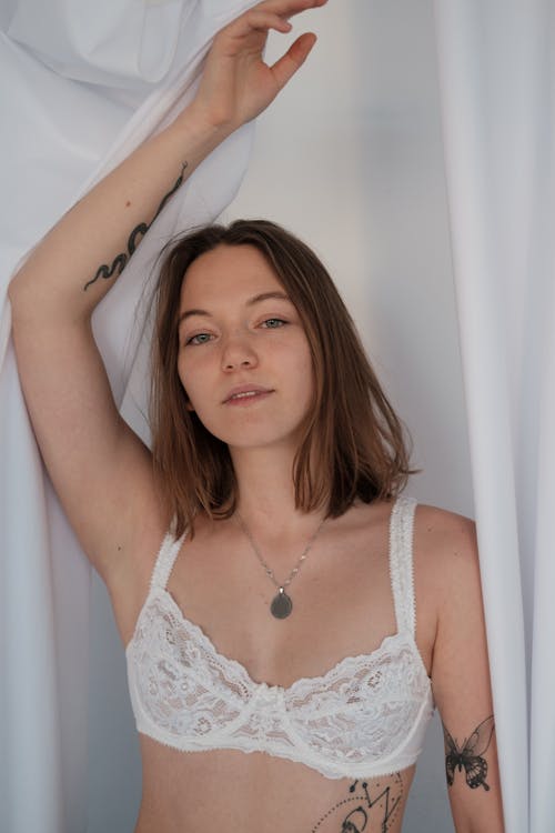 Free Confident young female with tattoos wearing white lingerie looking at camera with raised arm while standing near light tulle in studio Stock Photo