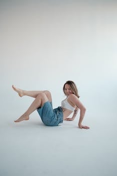 A cheerful young woman in denim shorts and a bra poses playfully on a white background.