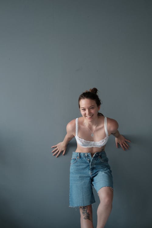 Joyful young female model wearing white underwear and denim shorts looking at camera while leaning on gray background in studio