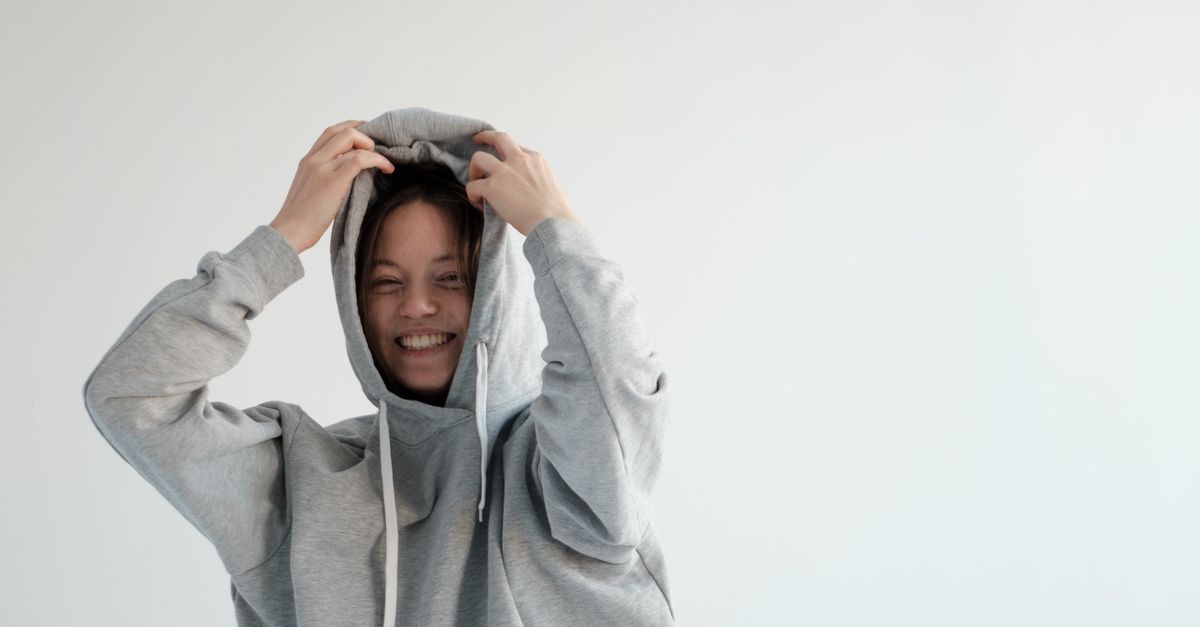 Happy young female putting on hood of trendy hoodie wearing denim shorts while standing on white background in light studio