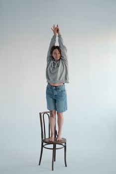 Full body of glad young female wearing casual hoodie and denim shorts looking at camera while standing on wooden chair with raised arms