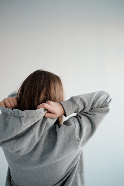 Back view of unrecognizable female with brown hair wearing gray hoodie while standing on white background in modern light studio
