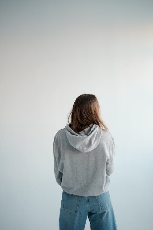 Back view of unrecognizable female with brown hair wearing gray hoodie and denim pants standing on white background in studio