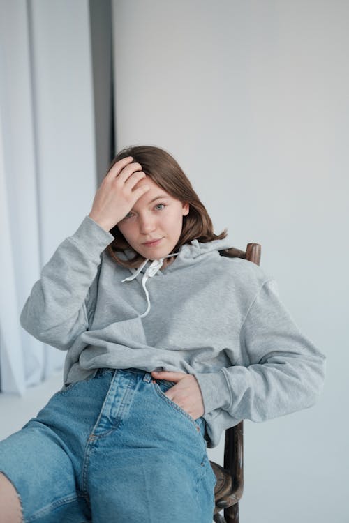 Free Serious female model wearing sweatshirt and denim shorts attaching hair while sitting on chair and looking at camera Stock Photo