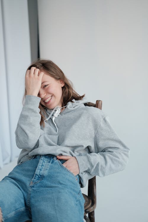 Positive young woman touching hair while laughing happily sitting on chair in studio and looking at camera