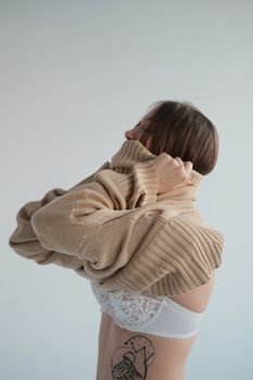 Side view of young female wearing underwear putting on soft beige sweater against gray background