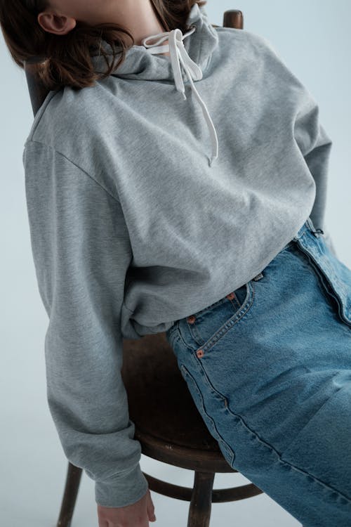 Free High angle of crop faceless female model wearing jeans and tied hoodie sitting on wooden chair in studio Stock Photo