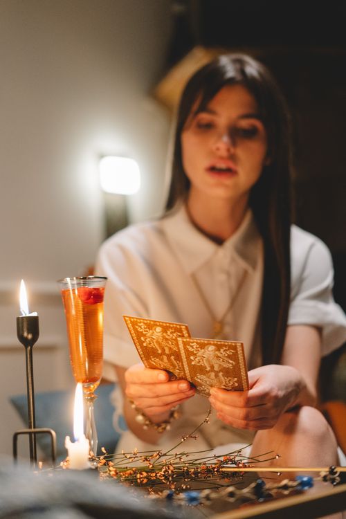 Free A woman reads tarot cards in a dimly lit room, with candles and a cocktail beside her, creating a mystical atmosphere. Stock Photo