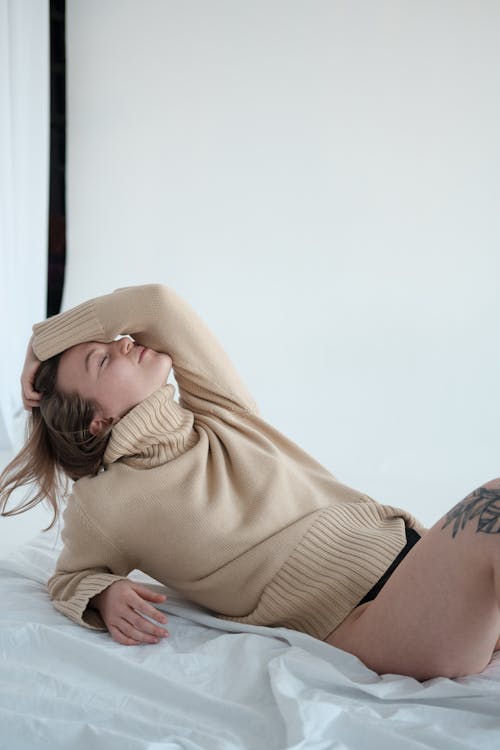 Side view of sensual female with closed eyes wearing underwear and sweater lying on bedsheet