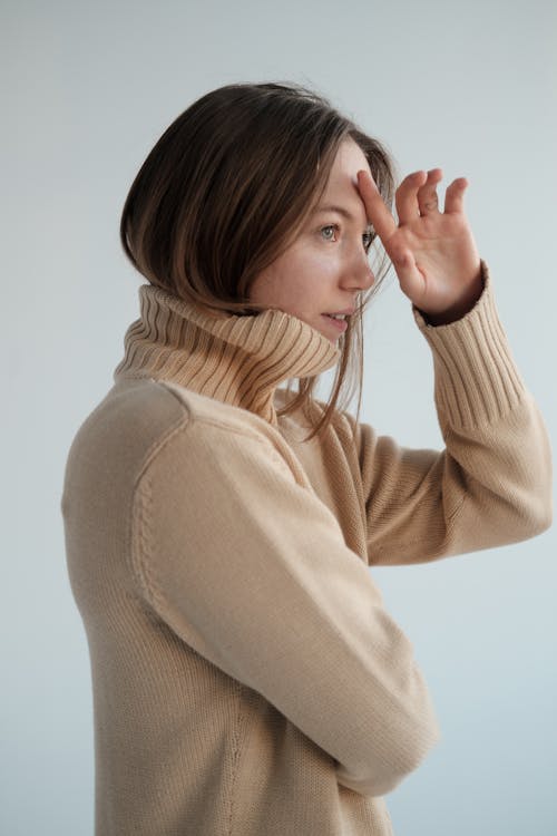 Free Side view of young female with long hair wearing warm sweater touching head while looking away against gray background Stock Photo