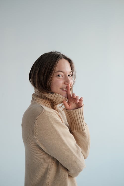 Free Side view of positive female wearing beige turtleneck covering mouth with finger against white background and looking at camera Stock Photo