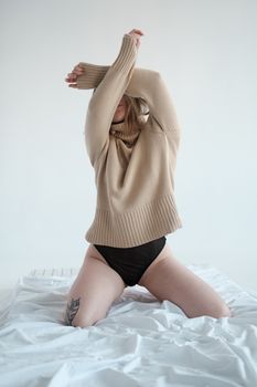 Young unrecognizable female in panties and sweater kneeling on crumpled bed and covering face with raised arms