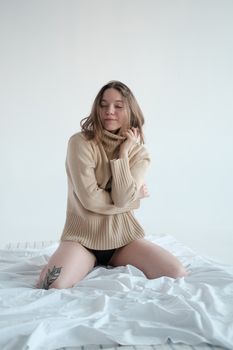Smiling female with closed eyes wearing underwear and turtleneck sitting on bed with white sheets against light background