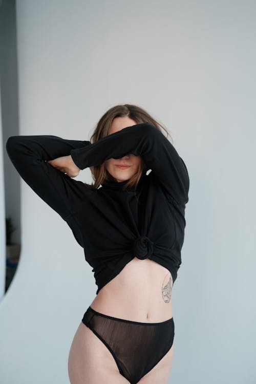 Graceful young female wearing black underwear and sweater covering eyes with arm and standing on white background in photo studio