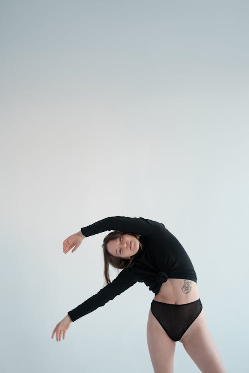 Attractive fit female wearing black panties and sweater bending to side with arms raised and looking at camera on white background