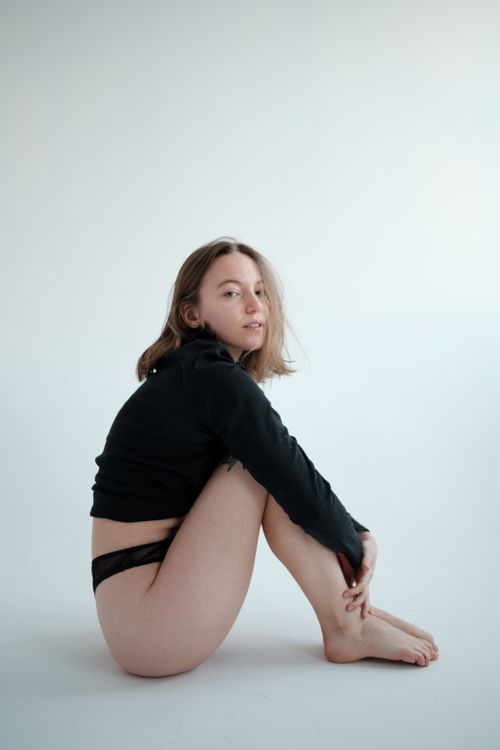 Free Full body side view of young female in panties and long sleeve top sitting against white background and looking at camera Stock Photo