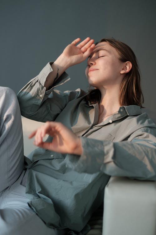 Free Positive female in linen shirt closing eyes and hiding face from bright sunshine while sitting on sofa Stock Photo