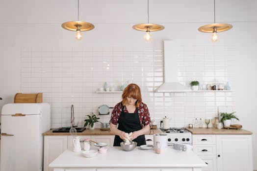 A woman wearing an apron prepares food in a stylish white kitchen.