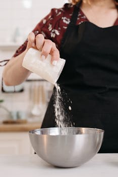 Woman in apron pouring flour into a bowl in kitchen, preparing food.