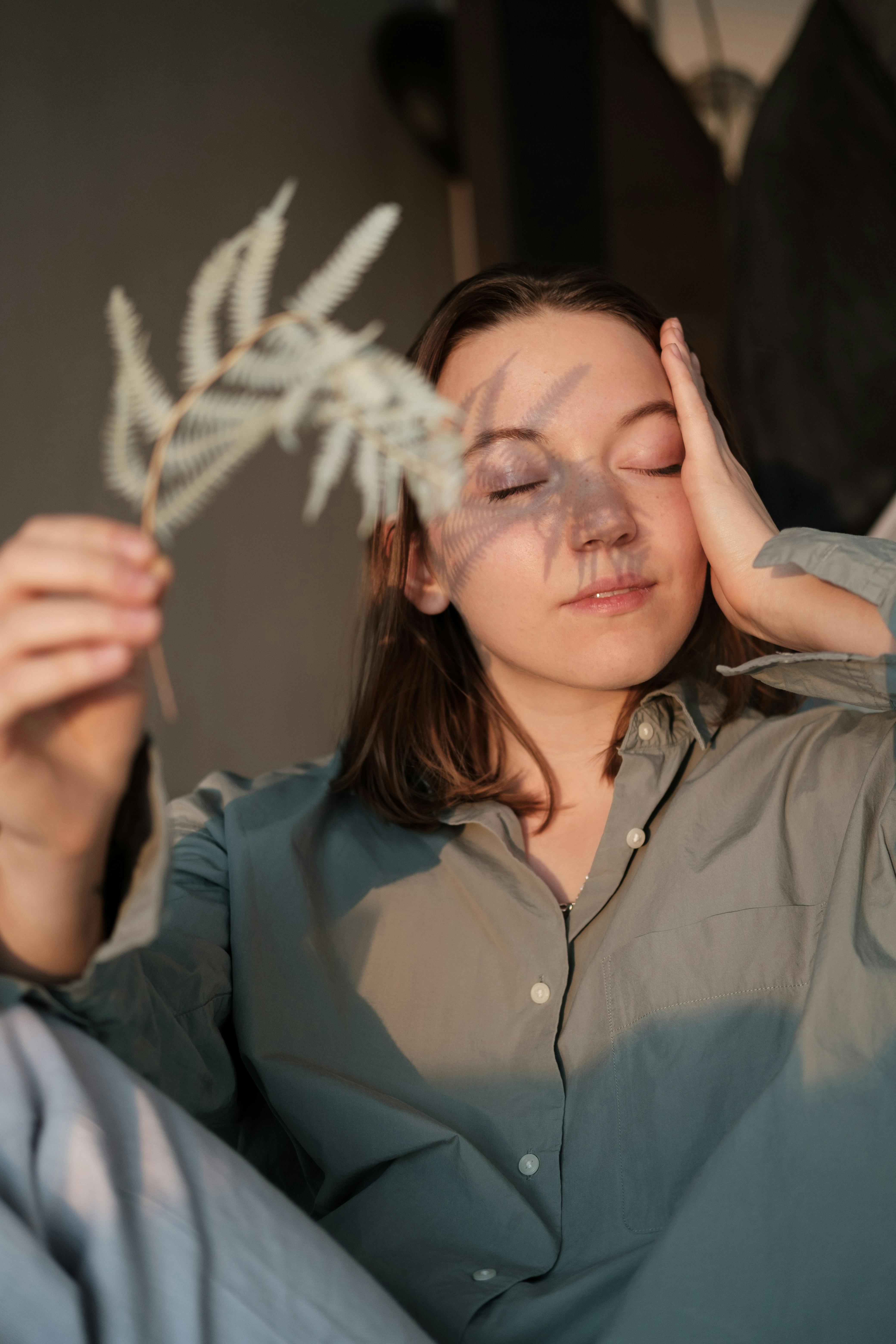 Delicate female with closed eyes touching face while hiding from sunlight with branch of fern