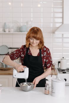Smiling woman blends ingredients in a modern kitchen using an electric mixer.