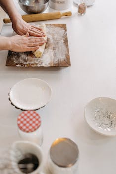 Hands kneading dough on a wooden board with baking tools on a white table.