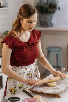 A woman in a floral apron rolls dough with a rolling pin on a kitchen countertop.