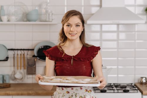 Free Smiling woman in kitchen holding tray of freshly baked cookies. Warm and inviting atmosphere. Stock Photo
