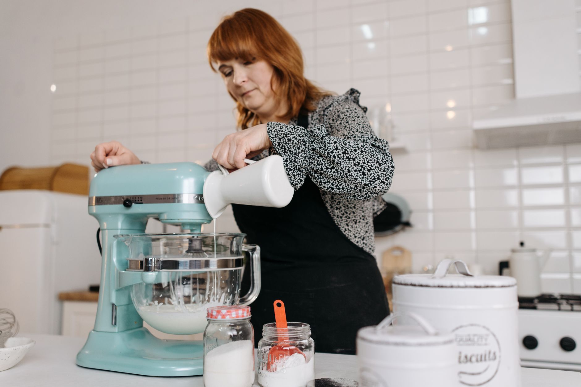 Woman pours ingredient into mixer bowl during food preparation in a modern kitchen setting.