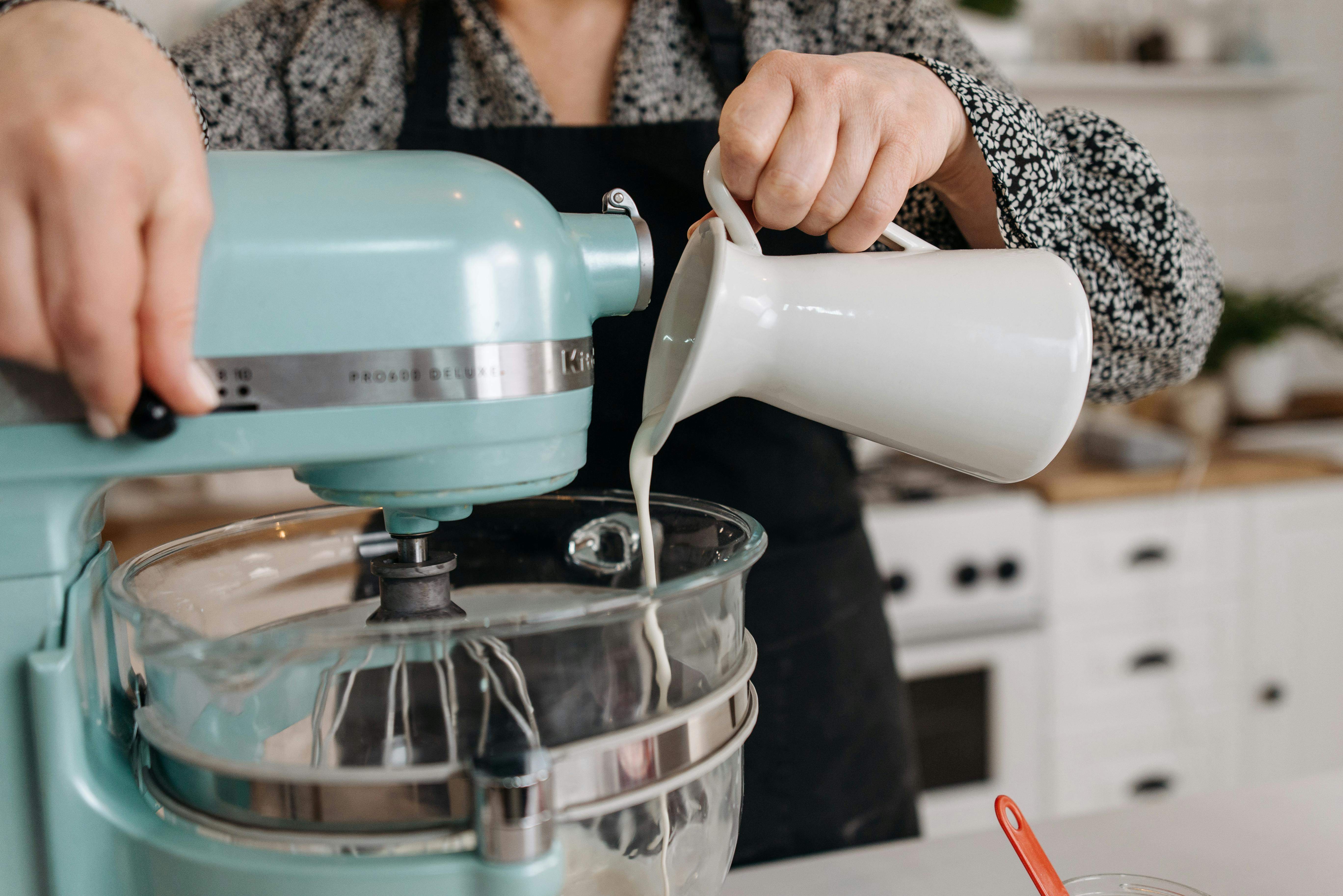 Person in kitchen pouring milk into a stand mixer bowl during baking preparation.