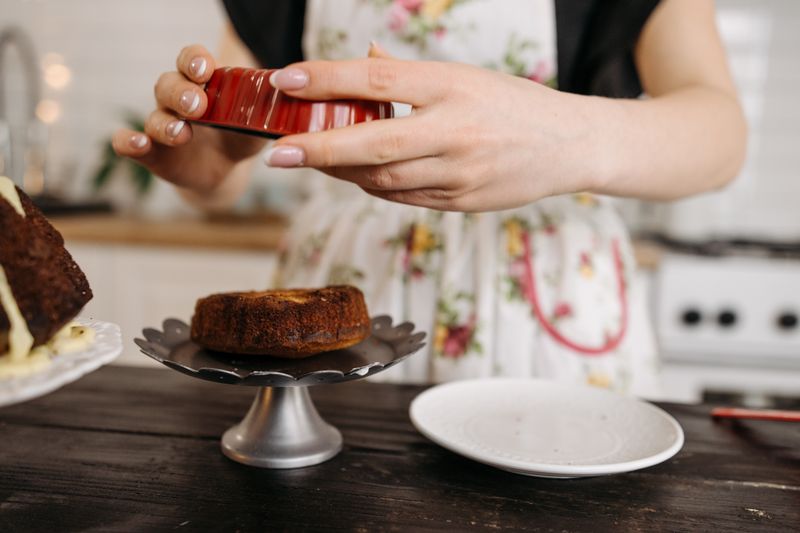 Close-up of hands demolding a small cake from a mold onto a cake stand in a cozy kitchen.