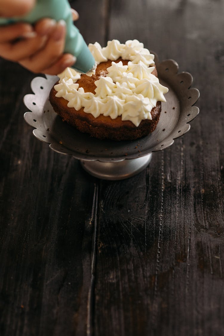 Person Piping White Icing On Brown Cake