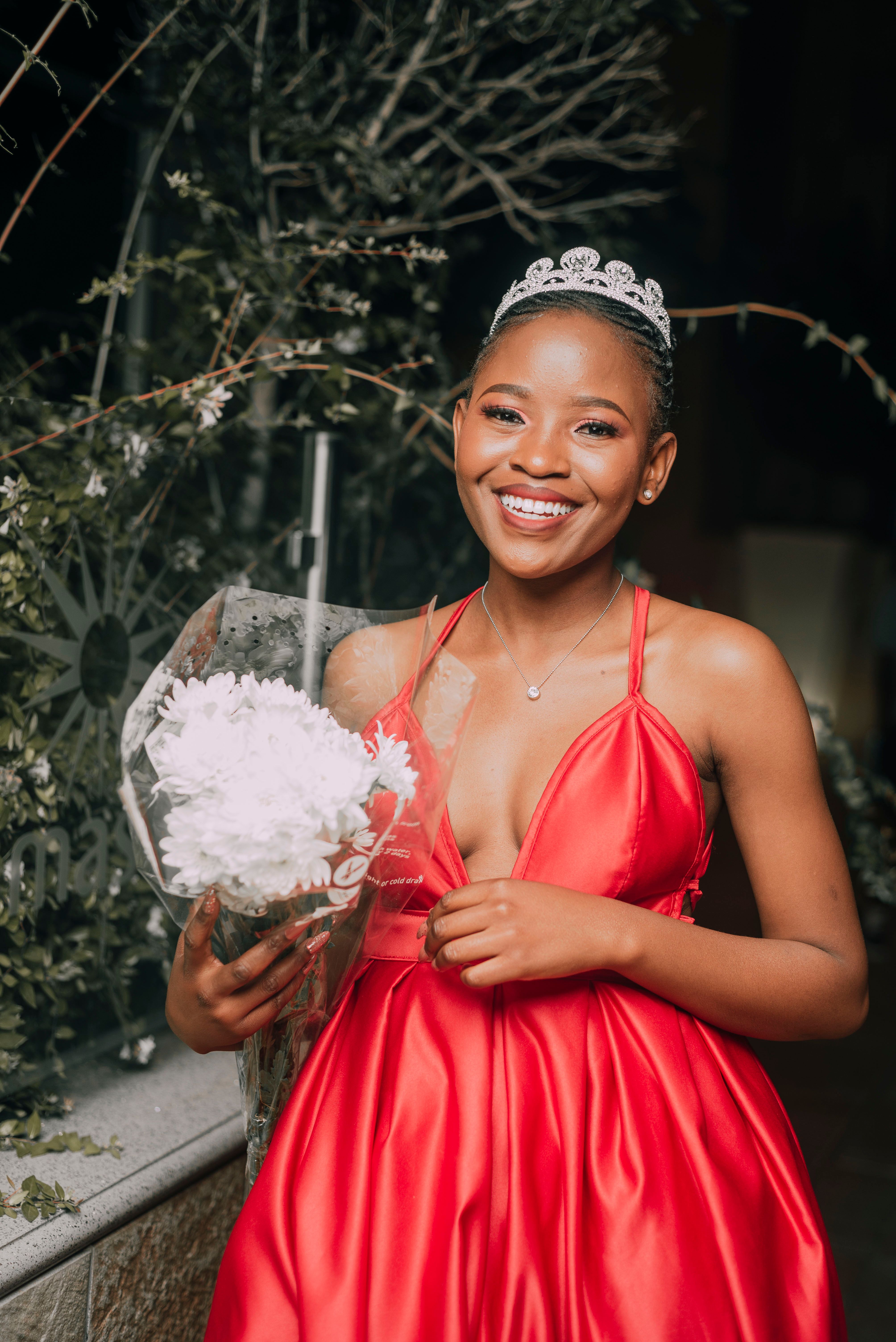 A woman in a red dress smiling while holding a bouquet, adorned with a tiara, creating a joyful portrait.