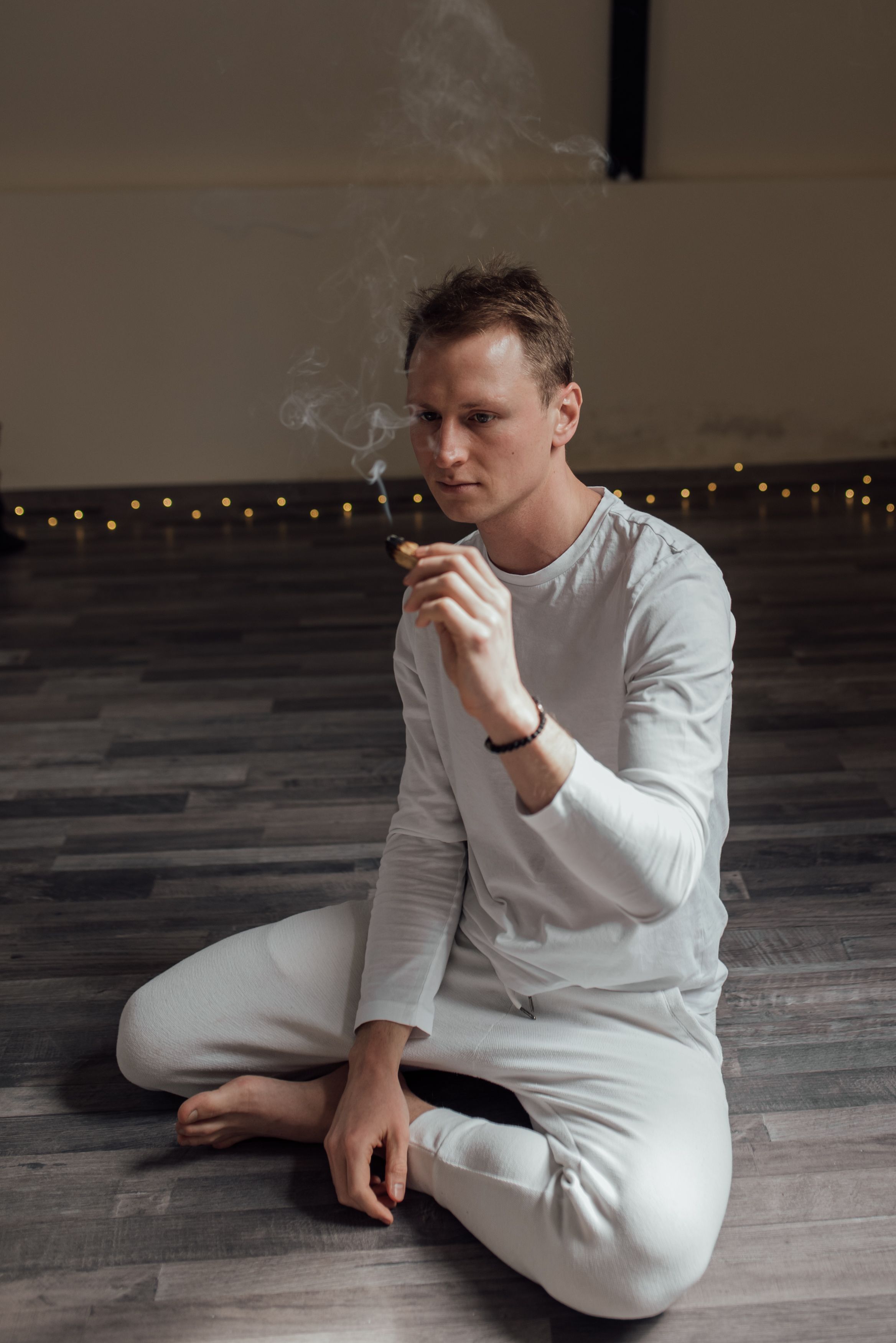 Man in white clothing meditates with palo santo, creating smoke for a calming spiritual atmosphere.