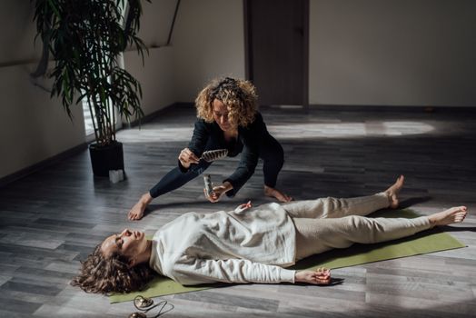 A Woman Holding a Feather and White Sage Beside a Woman Lying on Floor
