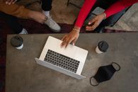 Top view of crop anonymous African American coworkers surfing netbook while sitting at table with medical mask and takeaway cups of coffee during work