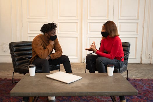 Focused African American colleagues in protective masks looking at each other while having conversation about work at table with laptop and paper cups of takeaway coffee