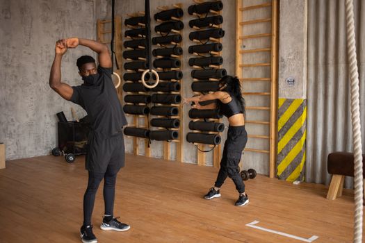 Sportive African American couple in activewear and medical masks doing physical exercises together during workout in gym with sport equipment