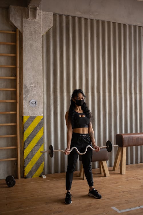 Young ethnic female in sportswear and protective mask lifting heavy barbell while training in gym