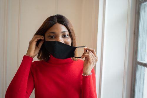 Free Young black female putting on protective mask looking at camera while standing against wall near window Stock Photo