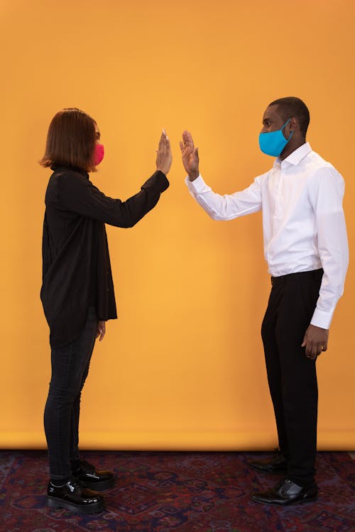 Full body of side view black friends in colorful medical masks joining hands at distance on yellow background