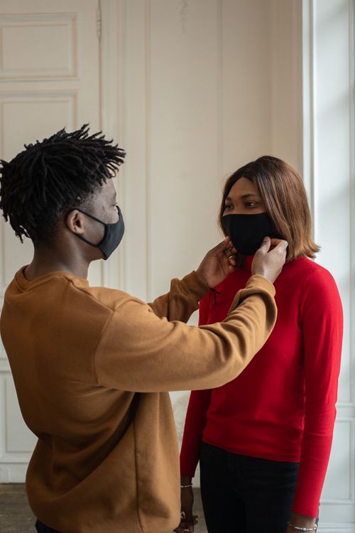 Free Young African American male putting protective mask on female friend face while standing together in light room and looking at each other Stock Photo