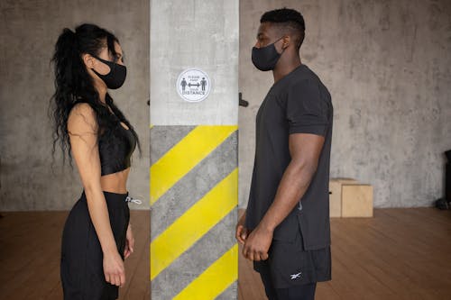 Side view young multiracial sportspeople in activewear and protective masks standing in gym and keeping social distance while looking at each other